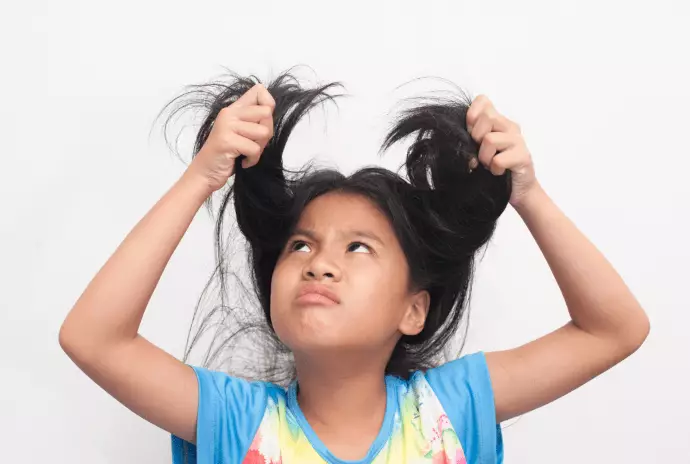 A photo of young girl with long black hair, holding strands of her hair above her head and frowning at it